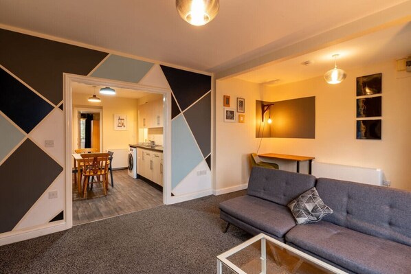 Una sala de estar moderna con un sofá gris y una mesa de centro de vidrio, un área de comedor con una mesa y sillas de madera, y una cocina visible a través de un umbral abierto.