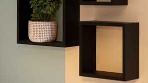 Een muur met drie zwarte, vierkante planken, waarvan er één een potplant bevat.