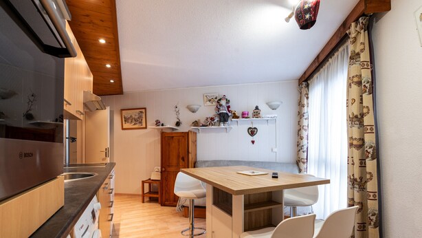 Una cocina compacta con un refrigerador Bosch, armarios de madera, una mesa de comedor de madera con dos sillas blancas y una decoración con forma de corazón en la pared.