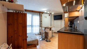 Habitación funcional con cama, kitchenette pequeña, zona de comedor con dos sillas y televisor montado en la pared.