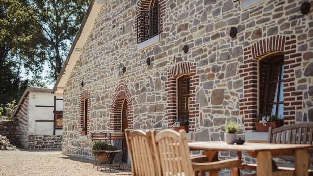 A stone building with arched windows and a courtyard with a wooden table and chairs. - La Bushaye - Cottage Le Studio (AUBEL)