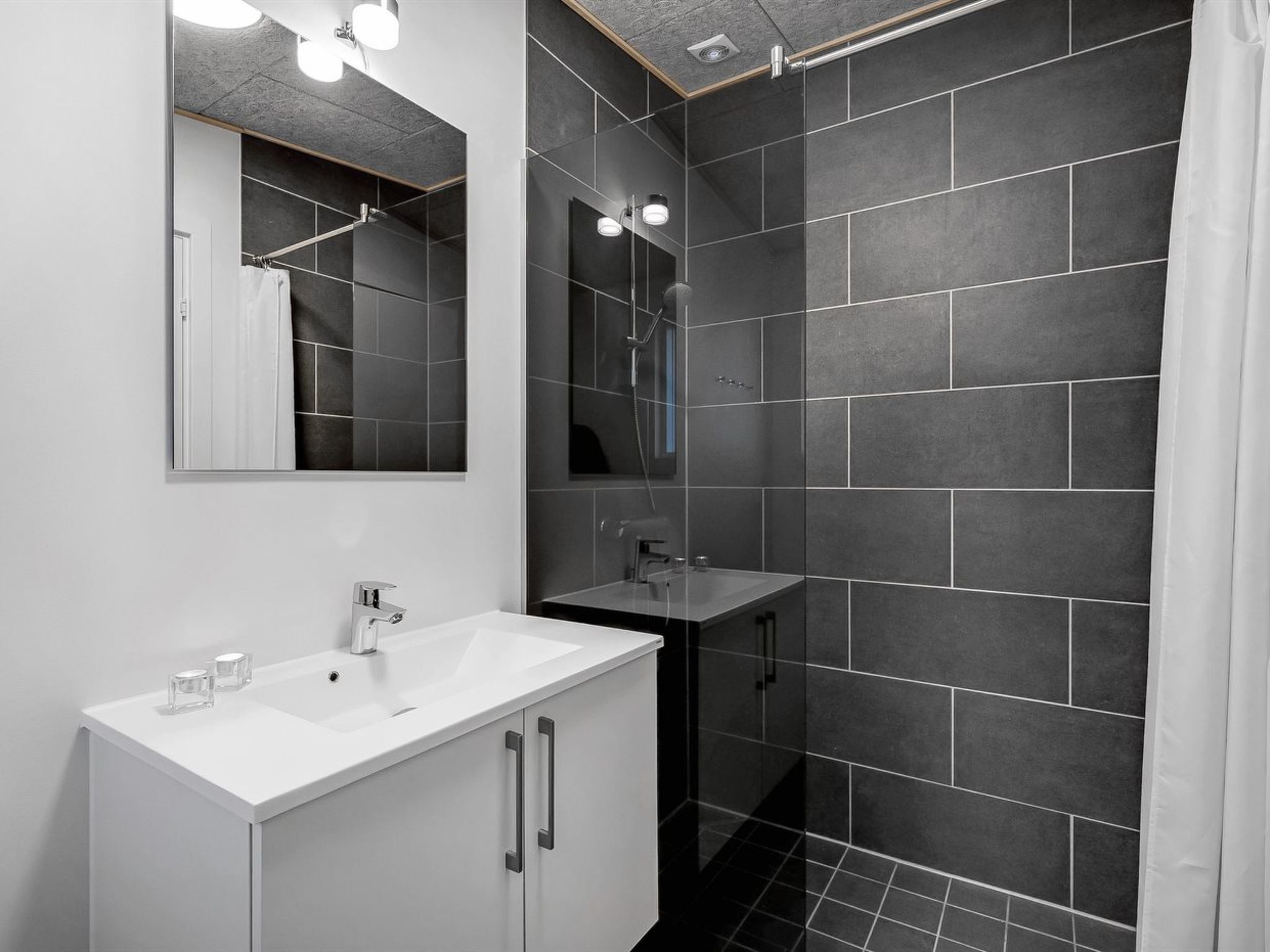 Un baño moderno con un lavamanos blanco, un espejo y una zona de ducha con azulejos oscuros.