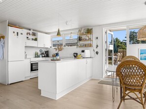 A modern kitchen with white cabinetry, a central island, and a dining area with a wicker chair. - "Zina" - 600m from the sea by Interhome (Hjørring)