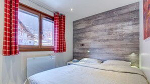 Un dormitorio con cabecera de madera, cama con ropa de cama blanca, mesita de noche, ventana con cortinas rojas y vistas a edificios.