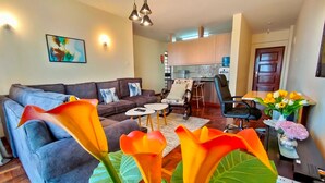 Ein modernes Wohnzimmer mit einer Couch, einem Couchtisch und Sesseln. Im Hintergrund ist eine Küchenzeile mit Schränken und einer Arbeitsplatte zu sehen.