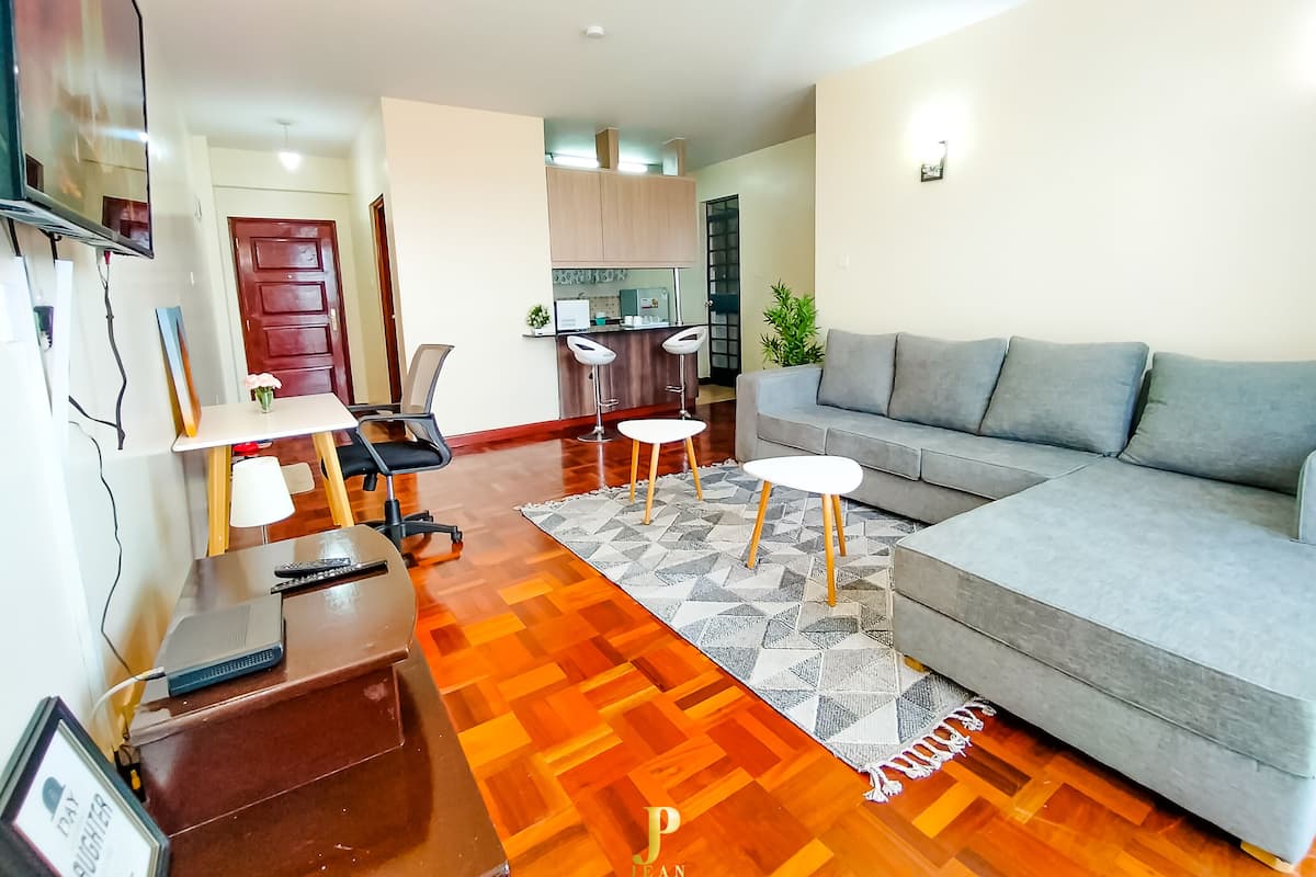 Een woonkamer met een bank, een salontafel, een televisie, een bureau met stoel, en een keuken in de achtergrond.