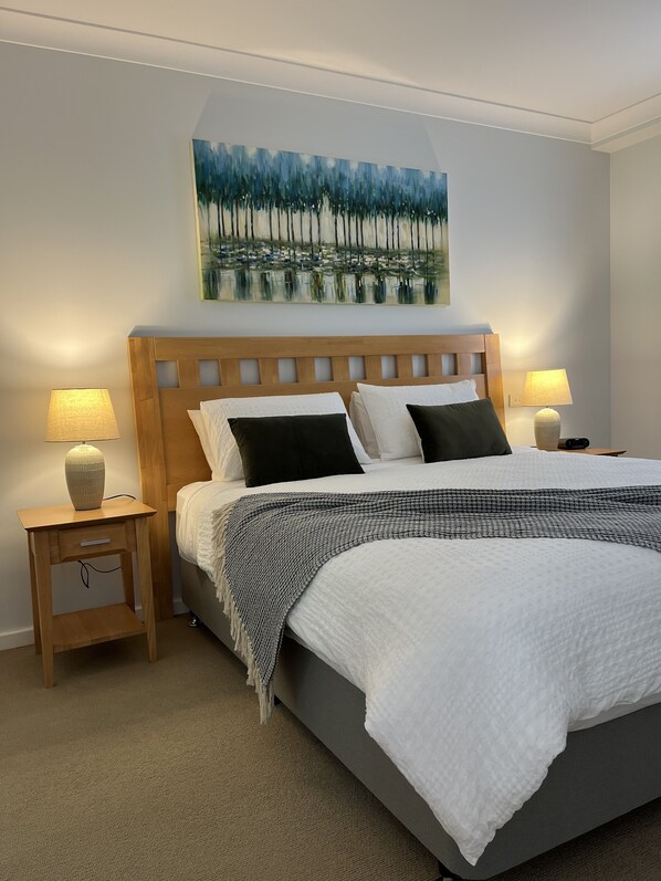 A neatly made bed with a wooden headboard, two bedside tables with lamps, a painting above the bed, and a carpeted floor. - City Escape Mounts Bay Apartment. Near Kings Park -  Parking - Wifi (Perth)