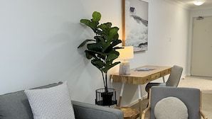 Een moderne woonkamer met een grijze bank, een houten bijzettafeltje, een grijze fauteuil, een zwarte vaas met plant, een houten bureau met lamp, en een lijst met foto aan de muur.