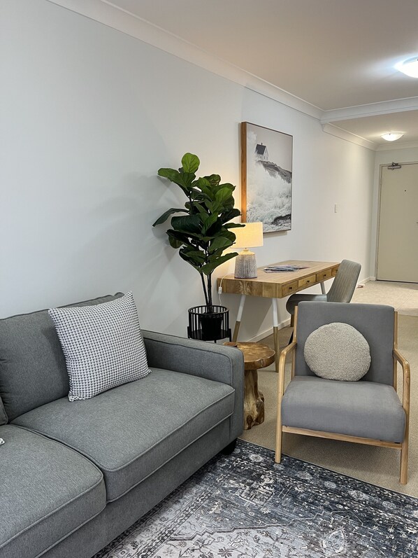 A modern living room with a grey sofa, a wooden side table, a grey armchair, a black vase with a plant, a wooden desk with a lamp, and a framed picture on the wall. - City Escape Mounts Bay Apartment. Near Kings Park -  Parking - Wifi (Perth)