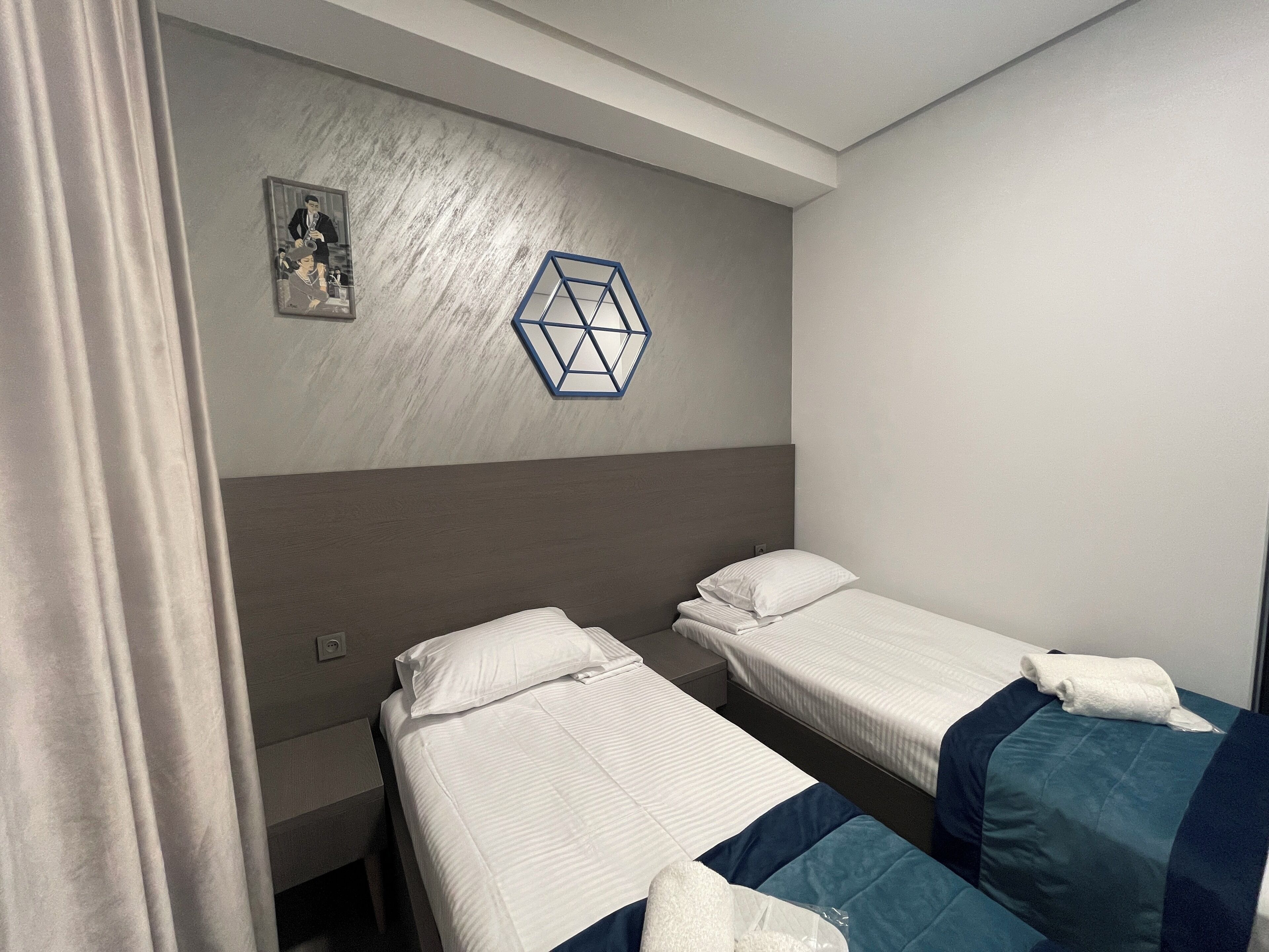 Une chambre d’hôtel avec deux lits simples, une table de chevet, un luminaire géométrique fixé au mur et un petit tableau encadré.