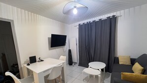 A modern living room with a flat-screen TV mounted on the wall, a dining table with chairs, a sofa with cushions, and a large window with curtains. - Ouréa, Appartement aux Portes de Ax les Thermes (Savignac-les-Ormeaux)