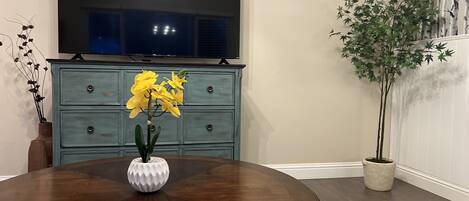 Un comedor con una mesa redonda de madera, un jarrón con flores amarillas, un aparador azul y un televisor montado en la pared.