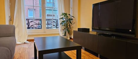 Ein Wohnzimmer mit einer Couch, einem Couchtisch, einem Fernseher auf einem Schrank, einer Topfpflanze und einem Fenster mit Blick auf Gebäude.