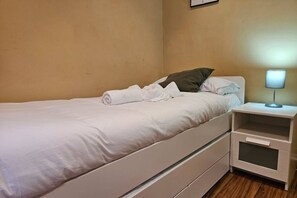 A single bed with a white mattress and pillows, a bedside table with a lamp, and a wall-mounted picture.