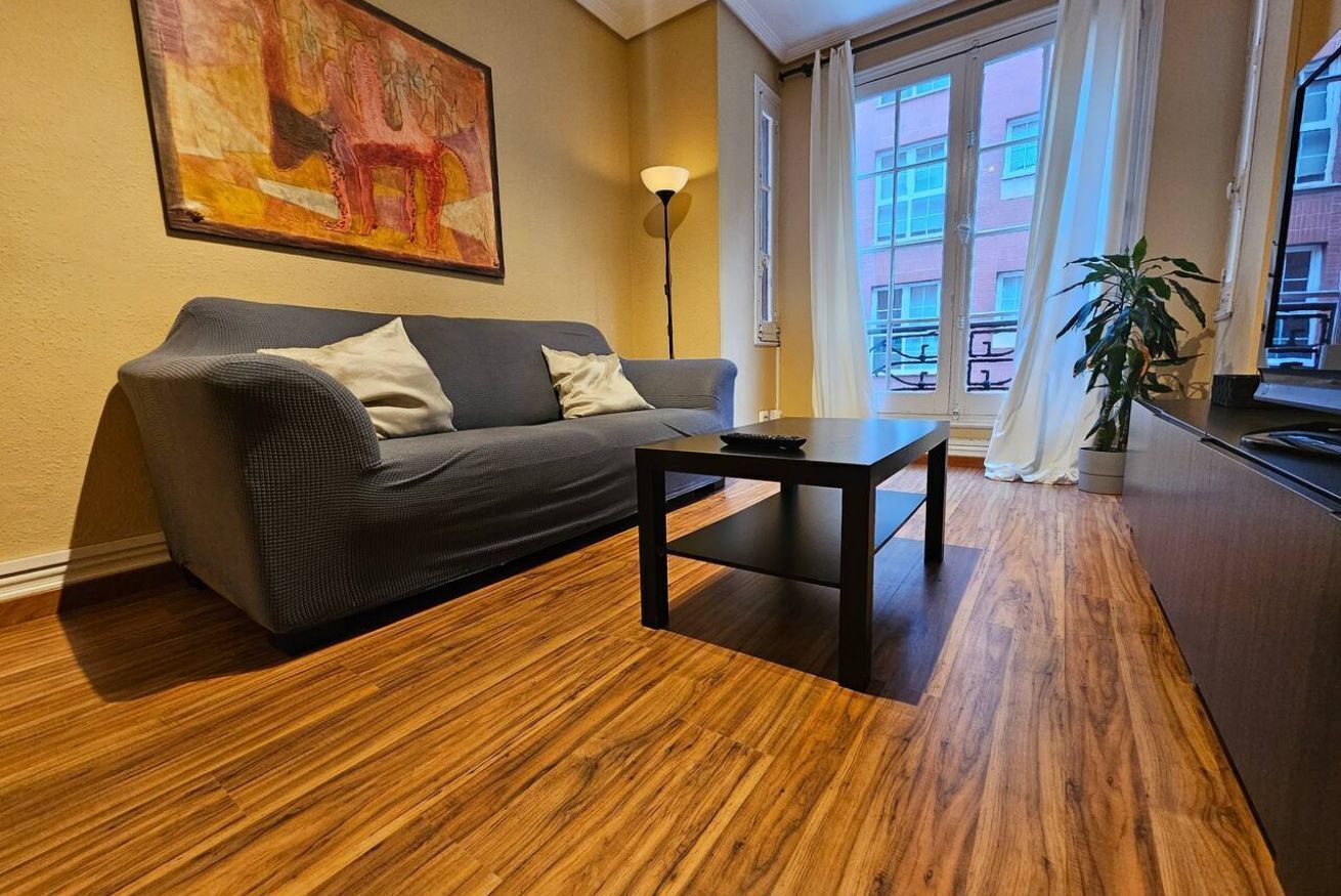 Een woonkamer met een bank, een salontafel, een schilderij aan de muur, een vloerlamp en een uitzicht op het omliggende landschap door grote ramen.