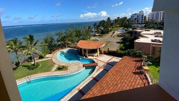 Vista de una piscina con diseño circular, rodeada de palmeras y una playa al fondo.