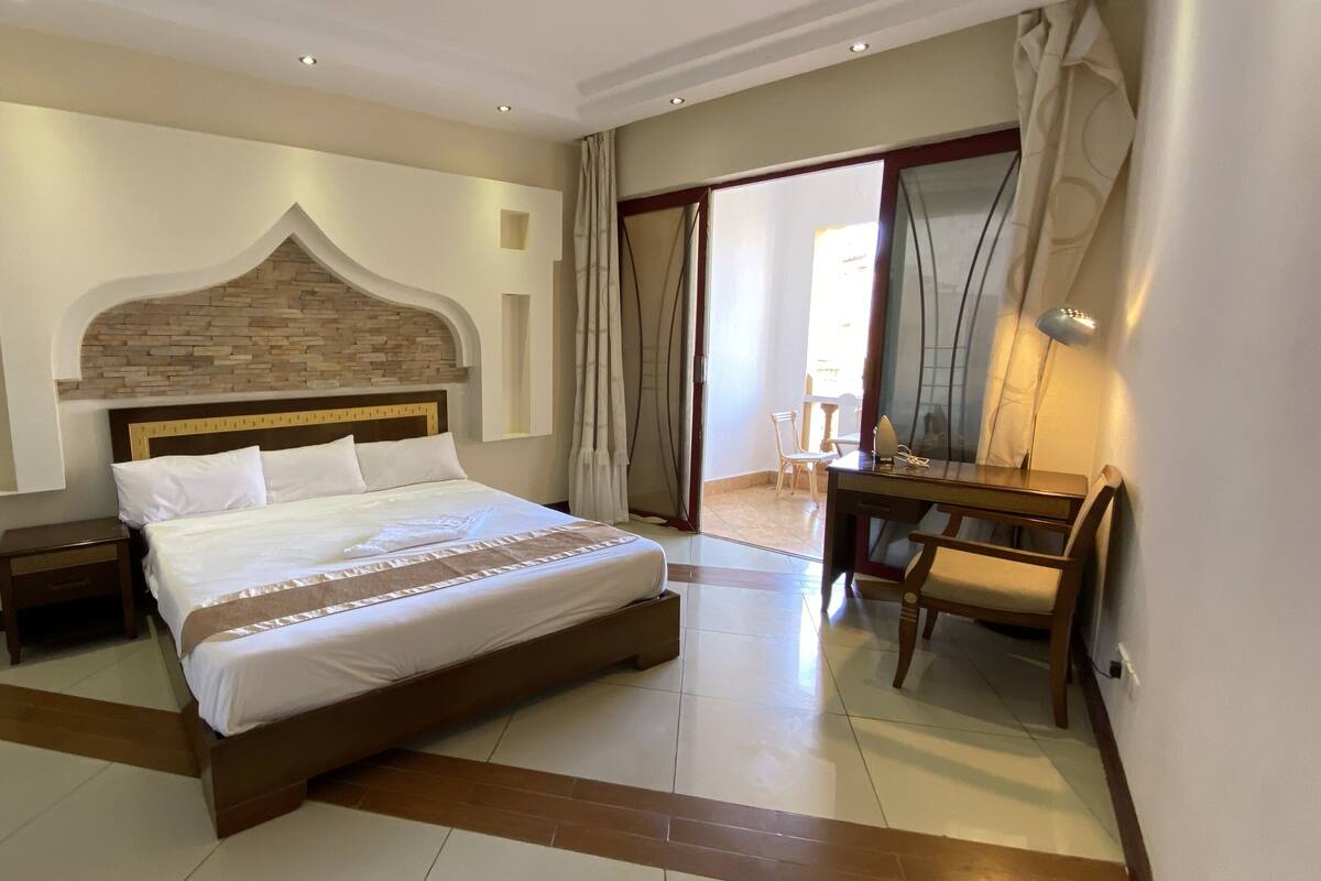 A hotel room with a large bed, a wooden headboard, a desk with a chair, and a large mirror.