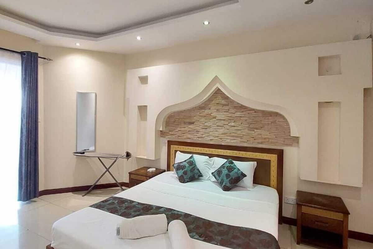 A hotel room with a large bed, a wooden headboard, a small bedside table, a mirror, and a window with curtains.
