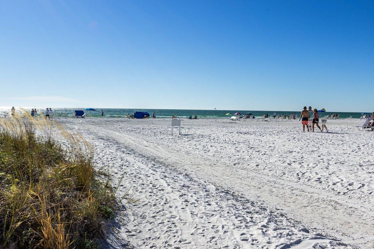A sandy beach with footprints, a clear sky, and a few people and umbrellas in the distance.