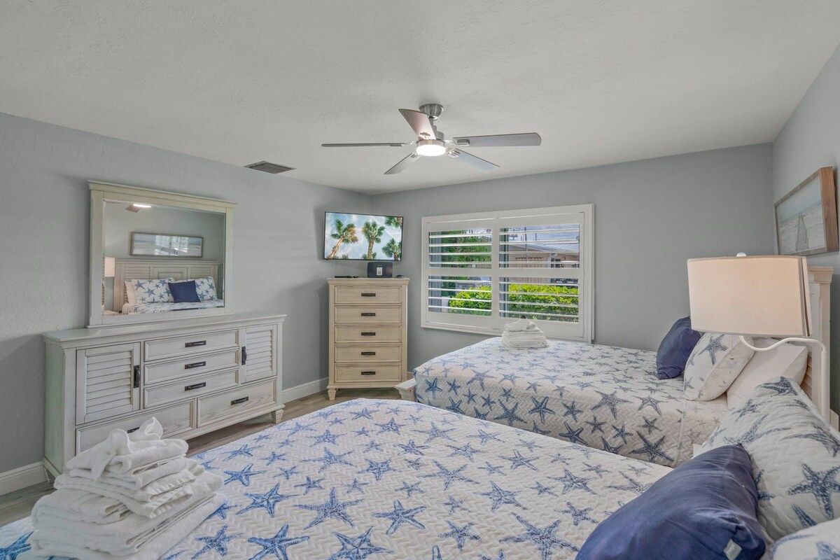 A bedroom with two beds, a dresser, a ceiling fan, a window with blinds, and a mirror.