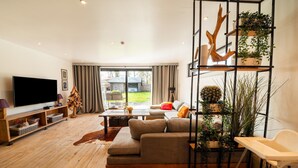 Una sala moderna con un televisor de pantalla plana grande, un sofá modular, una mesa de centro y una estantería. Se aprecia una vista del patio trasero a través de amplias ventanas.