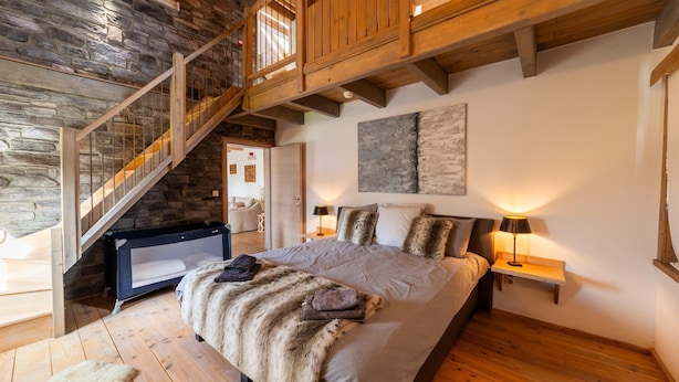 Un dormitorio con pared de piedra, escalera de madera, una cama grande con una manta de piel y una mesita de noche con lámpara.