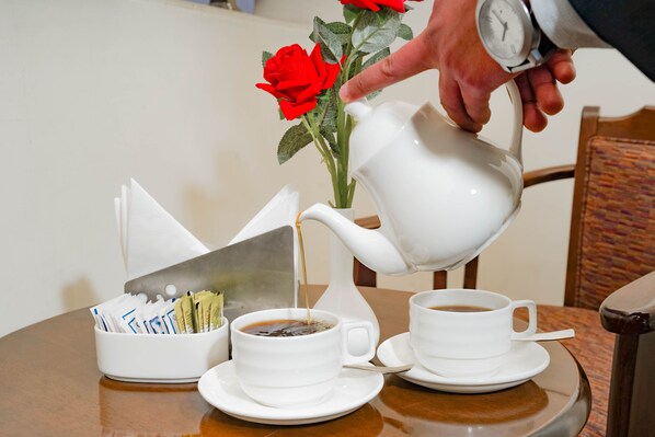 A white teapot pouring coffee into a cup, a vase with red roses, and a basket of papers on a wooden table.