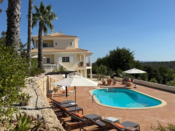 A poolside area with lounge chairs, umbrellas, and a view of a large house with a balcony. - Villa Botanica - Amazing villa with private pool (Silves)