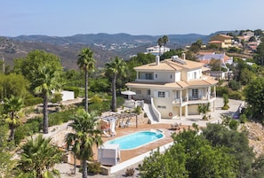 A large house with a swimming pool, surrounded by palm trees and a hilly landscape.