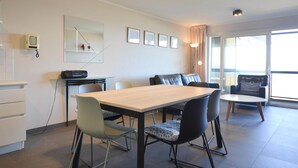 Un comedor moderno con una mesa de madera, sillas negras y grises, y un altavoz negro pequeño. Hay un espejo en la pared y una ventana con cortinas.
