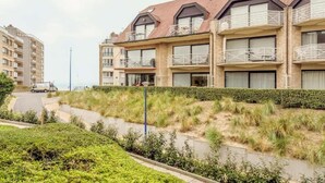 Un edificio residencial de varios pisos con balcones, un sendero pavimentado y un área verde con una fuente de agua.