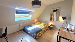 Un dormitorio con tragaluz, cama con ropa de cama estampada, mesita de noche con lámpara, espejo y escritorio con lámpara.