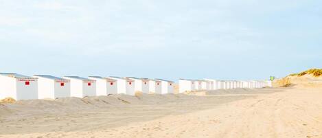 A row of white beach huts with red signage, arranged along a sandy beach.