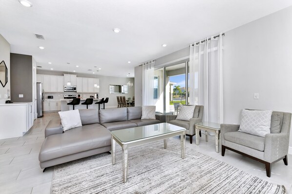 Un salon moderne avec un canapé gris, une table basse en verre et un fauteuil gris. On aperçoit à l’arrière-plan une cuisine avec des meubles blancs et un coin repas comprenant une table et des chaises.