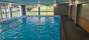 An indoor swimming pool with clear glass walls, a tiled floor, and a view of a grassy area outside. - Studio Dans Résidence Avec Piscine Couverte (Les Rousses)