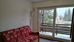 Una stanza con un divano in legno con un cuscino rosso a motivi a cuore, un'ampia finestra vista alberi e un edificio, e una ventola a soffitto.