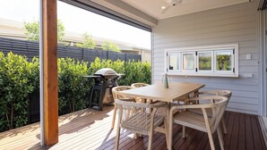 Eine Terrasse im Freien mit Holztisch, Korbstühlen, einem Grill und Blick auf einen Zaun und Grünflächen.