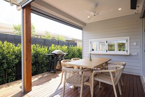 Eine Terrasse im Freien mit Holztisch, Korbstühlen, einem Grill und Blick auf einen Zaun und Grünflächen.