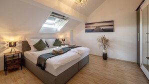 Une chambre avec une lucarne, un lit avec des oreillers, une table de chevet avec une lampe, un vase contenant des fleurs séchées et un tableau accroché au mur.