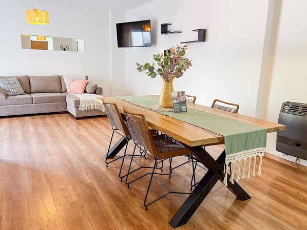 A modern living room with a wooden dining table, two chairs, a sofa, a TV, and a wall-mounted shelf.