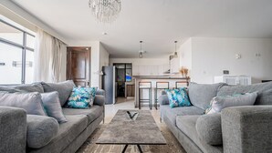 A modern living room with grey sofas and a coffee table, a kitchen in the background, and a large window with curtains. - Rose Upscale Apt (Nairobi)