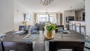 A modern dining area with a dark wooden table set for four, a vase of flowers, and a view of the kitchen and living area in the background. - Rose Upscale Apt (Nairobi)