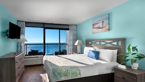 A bedroom with a large window, a bed, a dresser, a nightstand, and a TV.