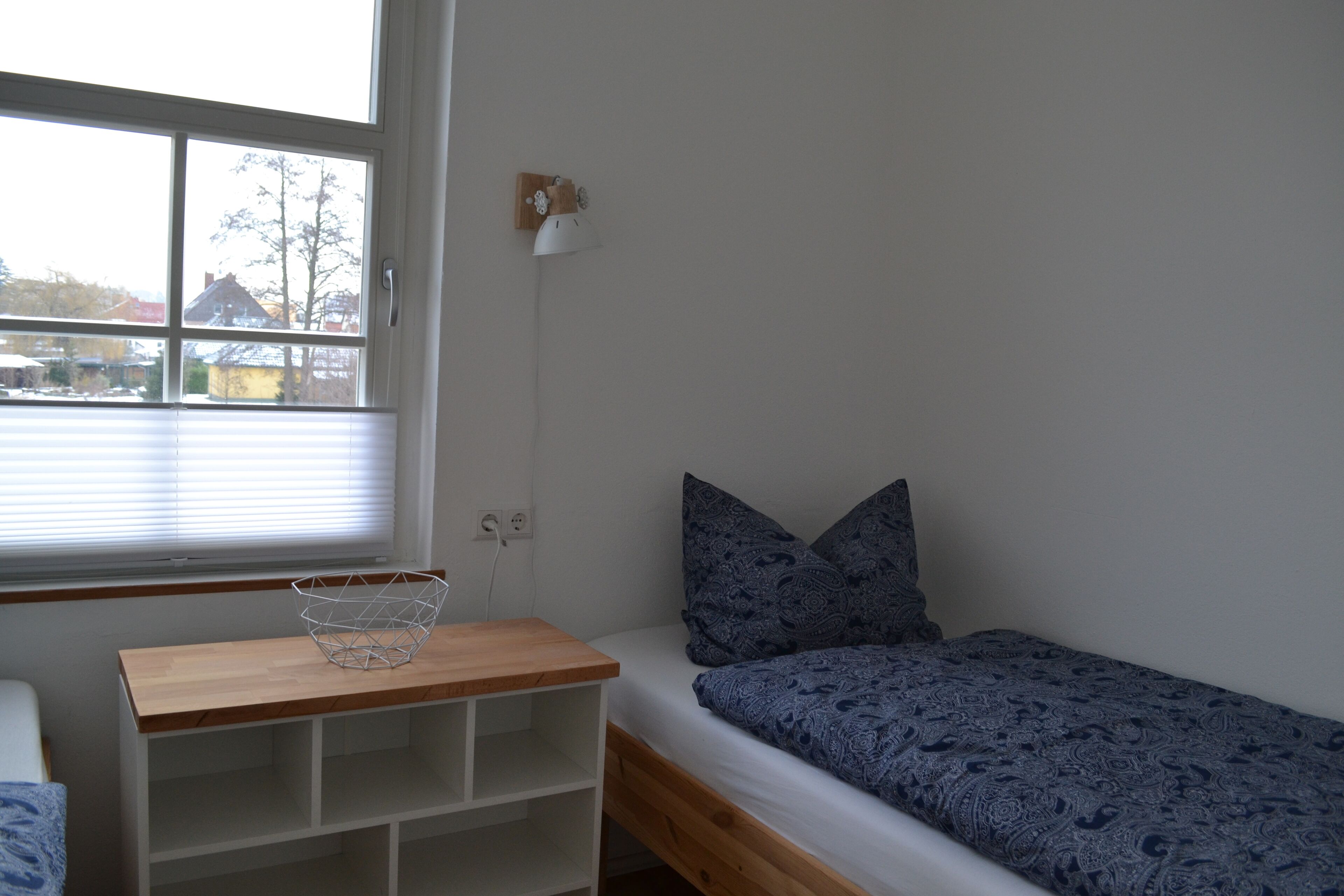 Ein kleines Zimmer mit einem Einzelbett, einem Holzkopfende, einem Nachttisch mit Drahtkorb und einem Fenster mit Jalousien.