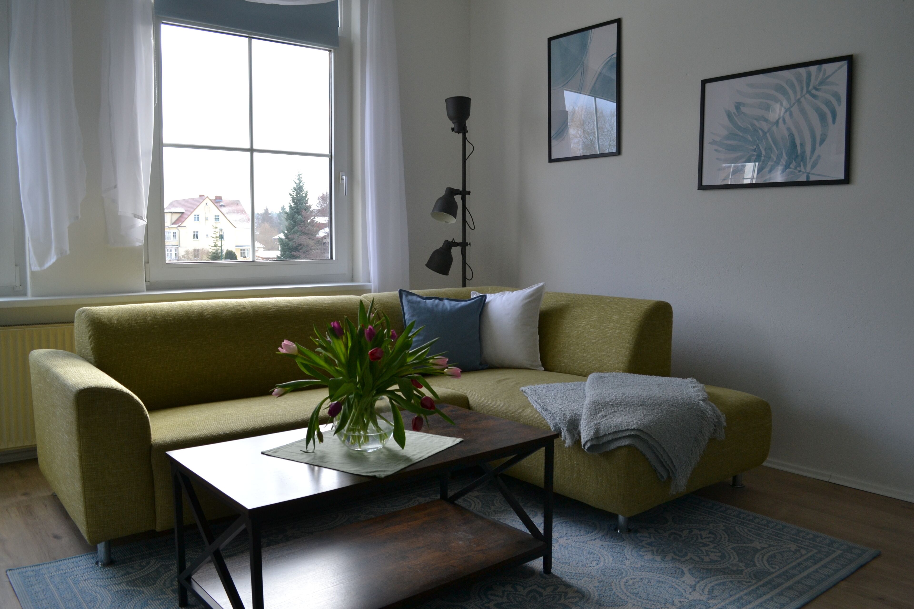 A living room with a sofa, a coffee table with a vase of flowers, a floor lamp, and a window with curtains.
