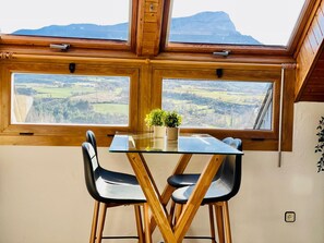 A dining area with a glass table and two chairs, a potted plant, and a view of a mountainous landscape through large windows. - Cozy 1-BR Apt with Pool, Near Downtown, Parking Included, Sleeps 4 (Jaca)
