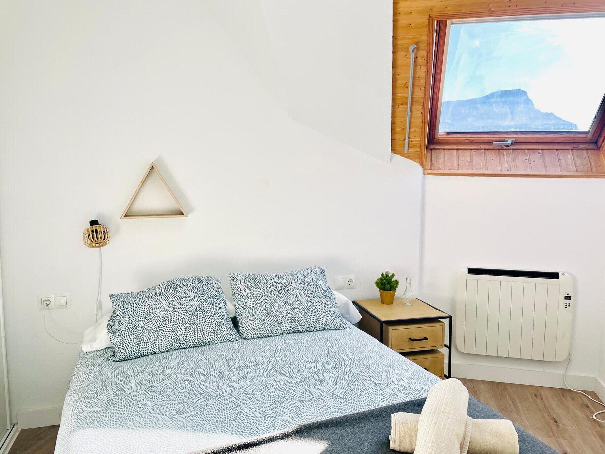 A bedroom with a bed, a bedside table, a radiator, and a window with a view of a mountain.