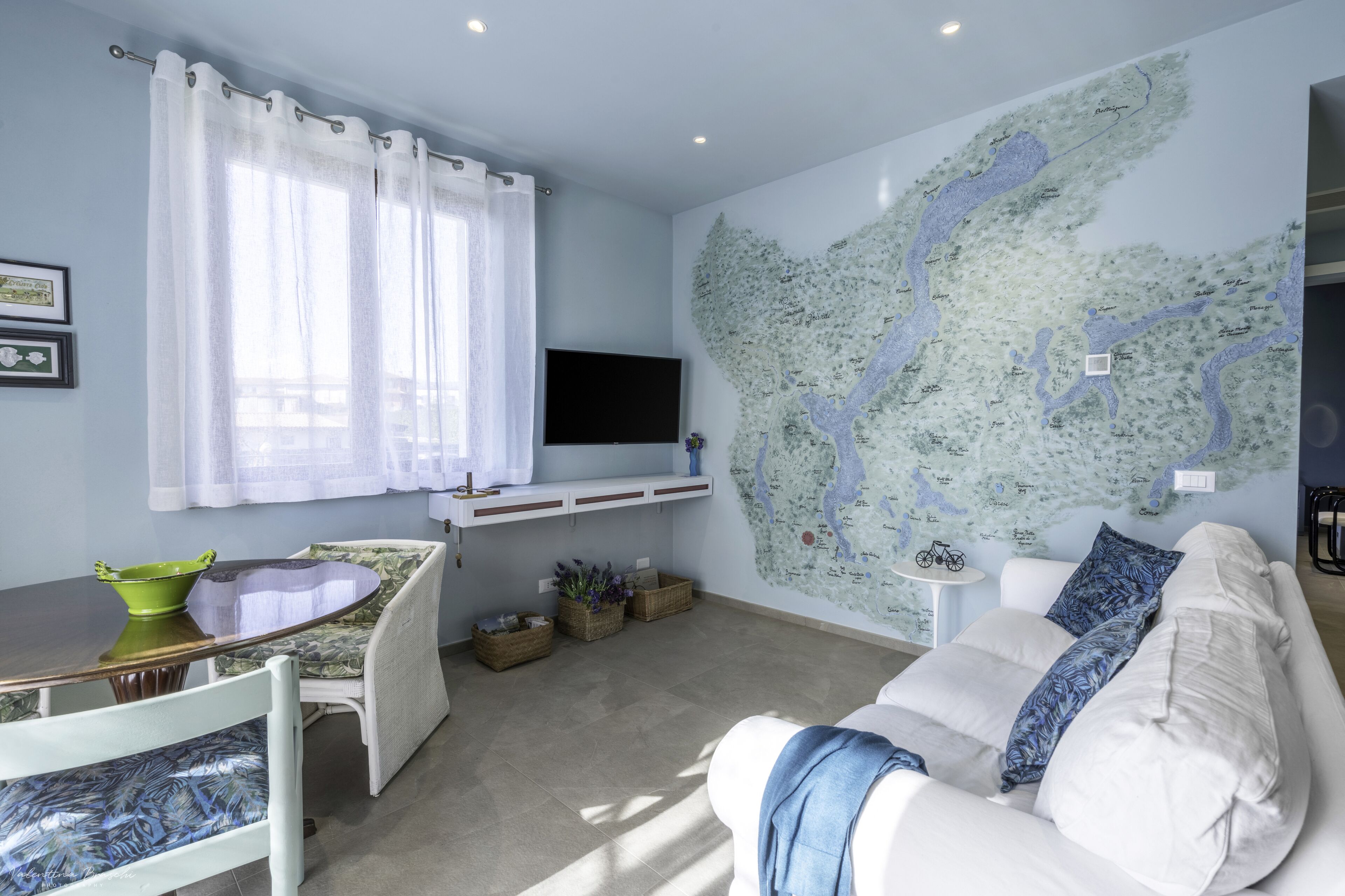 A living room with a white sofa, a dining table with chairs, a flat-screen TV mounted on the wall, and a large map on the wall.