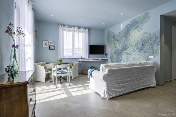 Un salon moderne avec un canapé blanc, une table à manger avec des chaises, un téléviseur fixé au mur et une fresque représentant une carte.