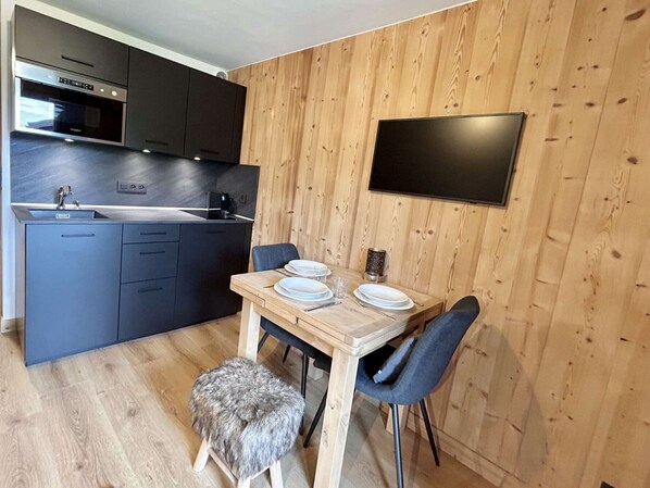 A modern kitchen with dark cabinets, a wooden dining table set for two, and a flat-screen TV mounted on the wall.